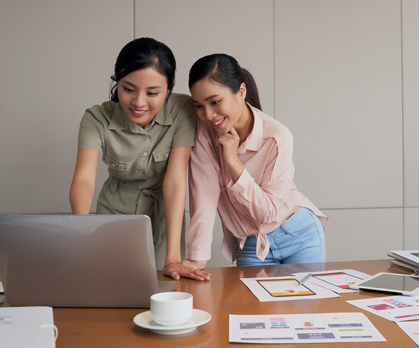Two young Asian female coworkers standing at a desk and using a laptop together